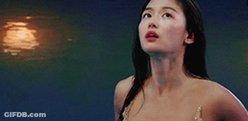 A Woman In A Mermaid Costume Is Standing In Front Of A Body Of Water Looking Up . GIF