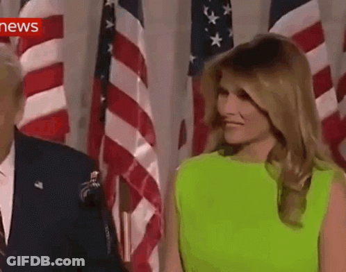 A Woman In A Neon Green Dress Is Standing Next To A Man In A Suit In Front Of American Flags . GIF