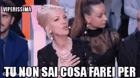 A Woman In A Pearl Necklace Is Sitting In Front Of A Group Of People With Her Hand On Her Chest . GIF