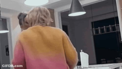A Woman In A Pink And Orange Sweater Is Standing In A Kitchen Looking At Herself In The Mirror . GIF