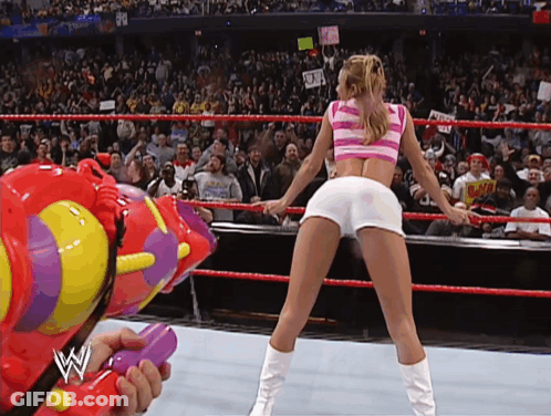 A Woman In A Pink And White Striped Top Is Dancing In A Wrestling Ring While A Man Holds A Water Gun GIF