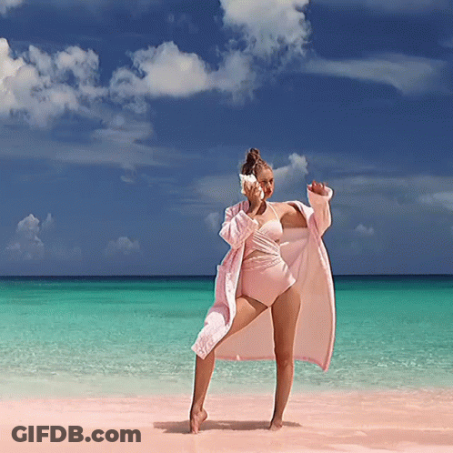 A Woman In A Pink Bathing Suit Is Standing On A Beach Holding A Seashell GIF