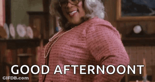 A Woman In A Pink Dress And Glasses Is Standing In A Room And Smiling . GIF
