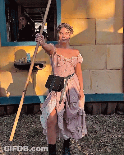 A Woman In A Pink Dress Holds A Wooden Stick GIF
