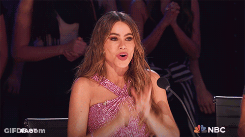A Woman In A Pink Dress Is Clapping In Front Of A Microphone With The Nbc Logo In The Background GIF