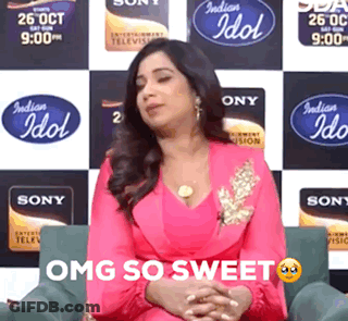 A Woman In A Pink Dress Is Sitting In Front Of A Wall With Indian Idol Logos On It GIF