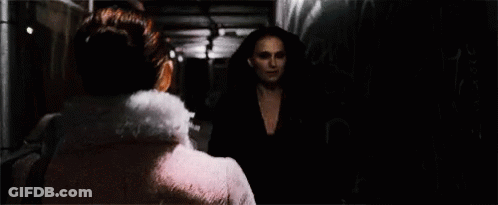 A Woman In A Pink Dress Is Standing In A Dark Room Looking At Another Woman . GIF