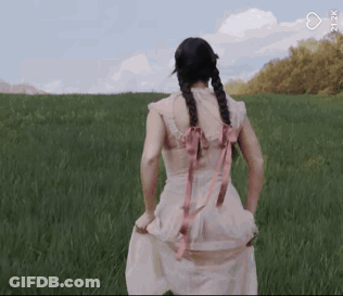 A Woman In A Pink Dress Is Standing In A Grassy Field With Her Hands In Her Pockets . GIF