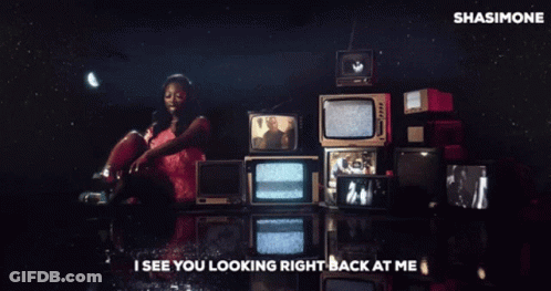 A Woman In A Pink Dress Is Standing In Front Of A Bunch Of Televisions With The Words I See You Looking Right Back GIF