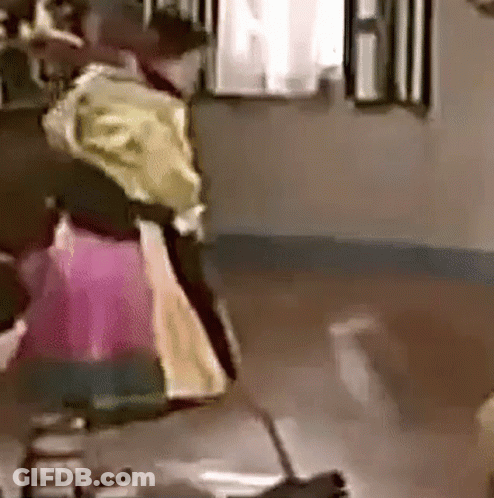 A Woman In A Pink Dress Is Standing On A Wooden Floor In A Room With A Broom . GIF
