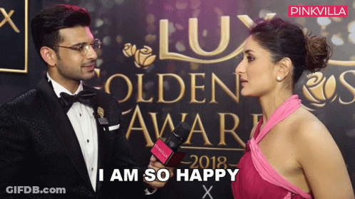 A Woman In A Pink Dress Is Talking To A Man In A Tuxedo In Front Of A Wall That Says Lux GIF