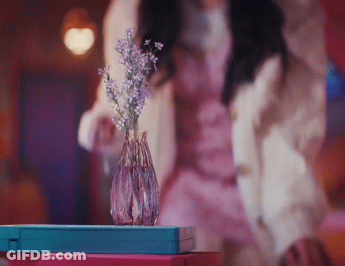 A Woman In A Pink Dress Stands In Front Of A Vase Of Flowers GIF
