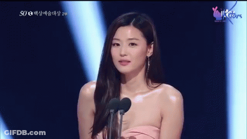 A Woman In A Pink Dress Stands In Front Of Two Microphones With A Heart On The Bottom Right Corner GIF