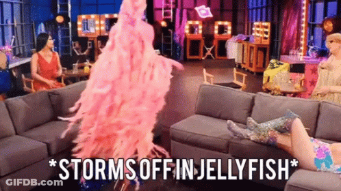 A Woman In A Pink Feathered Dress Is Standing In Front Of A Couch And Says Storms Off In Jellyfish . GIF