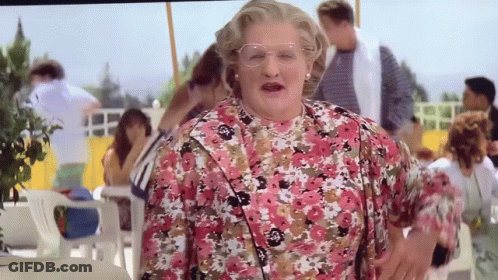 A Woman In A Pink Floral Dress And Glasses Stands In Front Of A Crowd Of People GIF