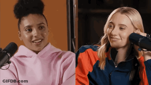 A Woman In A Pink Hoodie Is Talking Into A Microphone Next To A Woman In An Orange Jacket . GIF
