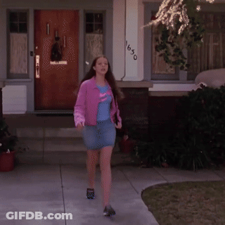 A Woman In A Pink Jacket And Blue Skirt Is Walking In Front Of A House With The Number 1650 On It GIF