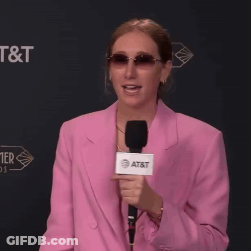 A Woman In A Pink Jacket Holds A Microphone That Says At & T On It GIF