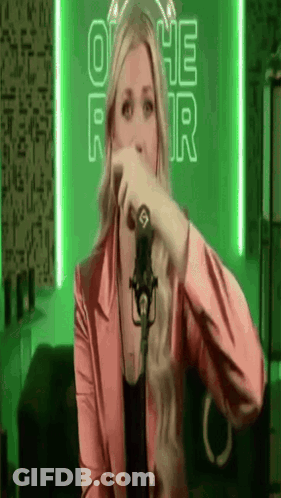 A Woman In A Pink Jacket Is Singing Into A Microphone In Front Of A Green Background . GIF