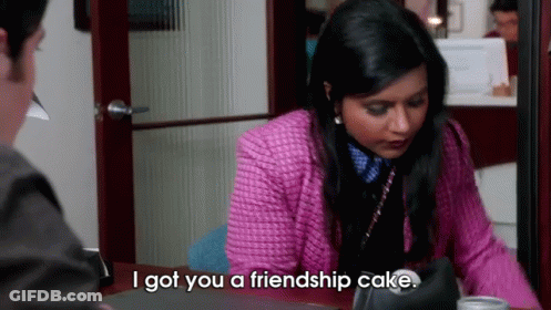A Woman In A Pink Jacket Is Sitting At A Desk Talking To A Man And Saying I Got You A Friendship Cake GIF