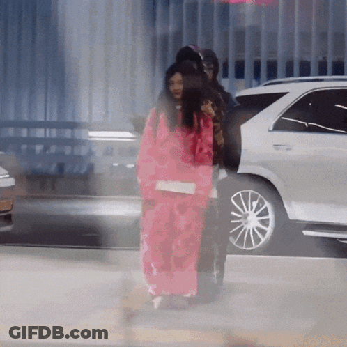 A Woman In A Pink Jacket Is Walking In Front Of A Car GIF