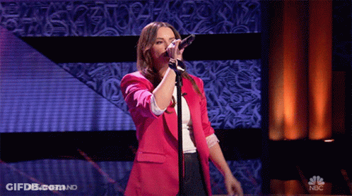 A Woman In A Pink Jacket Sings Into A Microphone On A Stage With A Nbc Logo Behind Her GIF