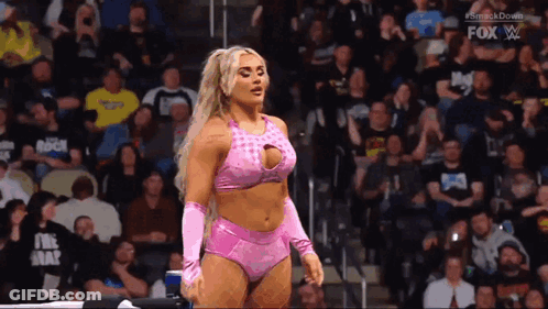 A Woman In A Pink Outfit Is Standing In Front Of A Crowd In A Wrestling Ring . GIF