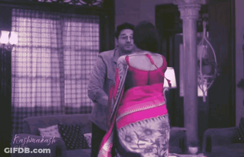 A Woman In A Pink Saree Is Standing Next To A Man In A Grey Jacket GIF