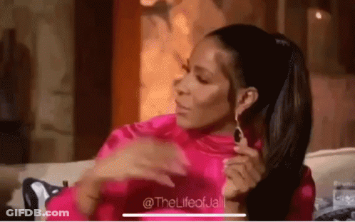 A Woman In A Pink Shirt And Earrings Is Sitting On A Couch With Her Hands On Her Chest . GIF