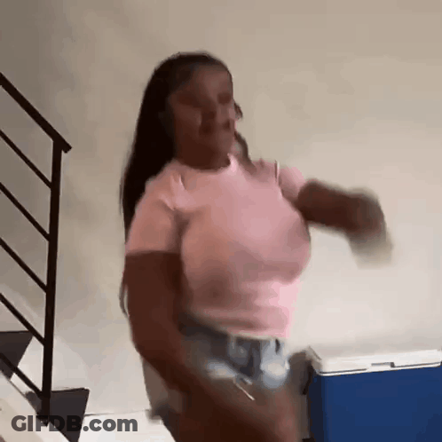 A Woman In A Pink Shirt And Shorts Is Dancing In Front Of A Blue Cooler In A Room . GIF