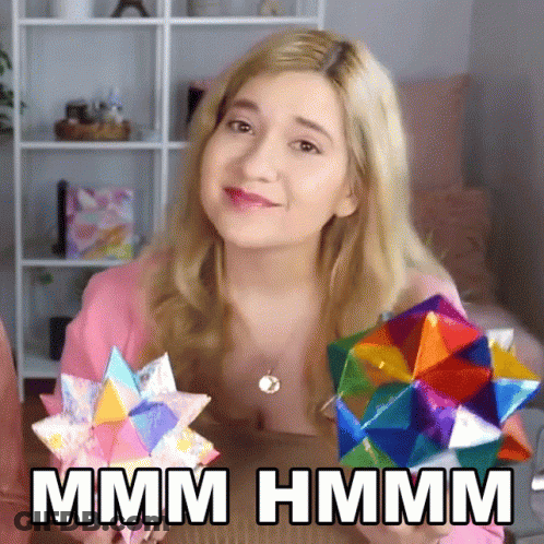 A Woman In A Pink Shirt Is Holding A Colorful Origami Object And Says Hmm GIF