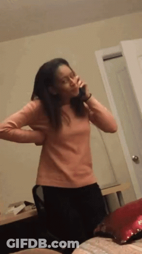A Woman In A Pink Shirt Is Standing In A Room Talking On A Phone GIF