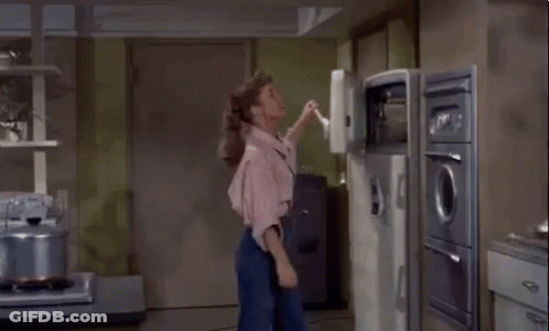 A Woman In A Pink Shirt Is Standing In Front Of An Open Refrigerator In A Kitchen GIF