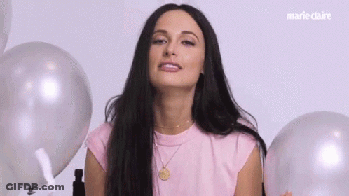 A Woman In A Pink Shirt Is Surrounded By Balloons And Making A Funny Face . GIF