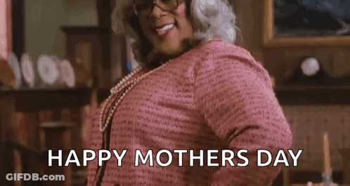 A Woman In A Pink Sweater And Glasses Is Standing In A Living Room And Smiling . GIF