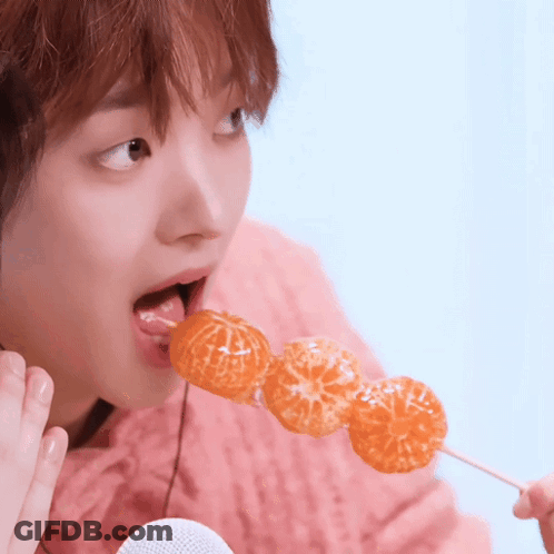 A Woman In A Pink Sweater Is Eating A Skewer Of Oranges On A Stick GIF