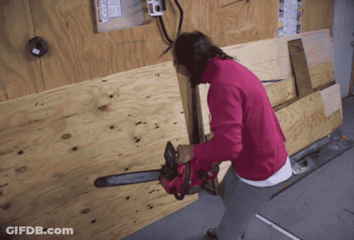 A Woman In A Pink Sweater Is Holding A Chainsaw Against A Wooden Wall GIF
