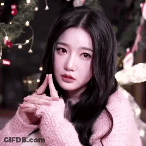 A Woman In A Pink Sweater Is Sitting In Front Of A Christmas Tree With Her Hands Folded . GIF