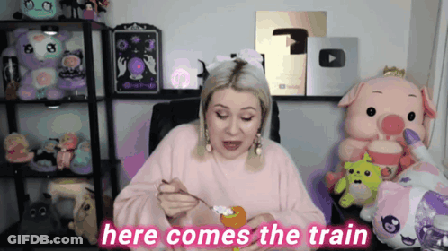 A Woman In A Pink Sweater Is Sitting In Front Of Stuffed Animals And Says " Here Comes The Train " GIF