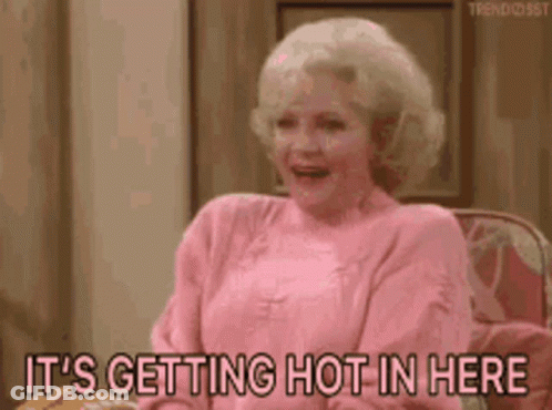 A Woman In A Pink Sweater Is Sitting On A Couch And Says It 'S Getting Hot In Here . GIF