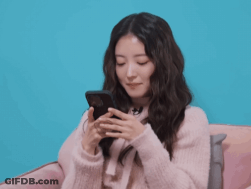 A Woman In A Pink Sweater Is Smiling While Holding A Cell Phone GIF