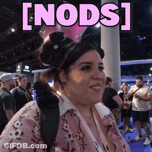 A Woman In A Pink Sweater Stands In Front Of A Sign That Says ' Nods ' GIF