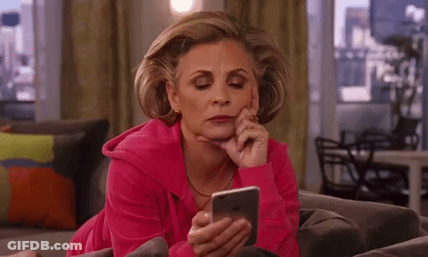 A Woman In A Pink Sweatshirt Is Laying On A Couch Looking At Her Phone . GIF