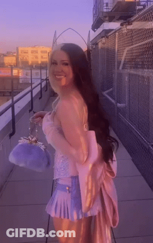 A Woman In A Pink Top And Blue Skirt Is Standing On A Balcony Holding A Purple Purse . GIF