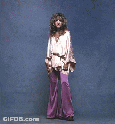 A Woman In A Pink Top And Purple Pants Stands In Front Of A Blue Background GIF