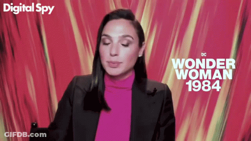 A Woman In A Pink Turtleneck And Black Jacket Is Sitting In Front Of A Digital Spy Wonder Woman 1984 Poster GIF