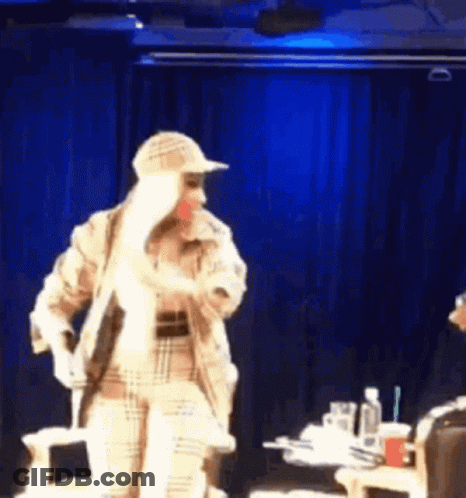 A Woman In A Plaid Coat And Hat Is Standing On A Stage . GIF