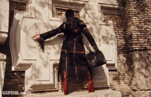 A Woman In A Plaid Coat And Red Boots Is Standing In Front Of A Brick Building GIF