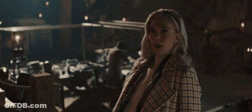 A Woman In A Plaid Coat Stands In A Dark Room Looking At The Camera GIF