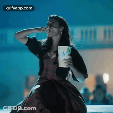 A Woman In A Plaid Dress Is Eating Popcorn And Holding A Cup . GIF